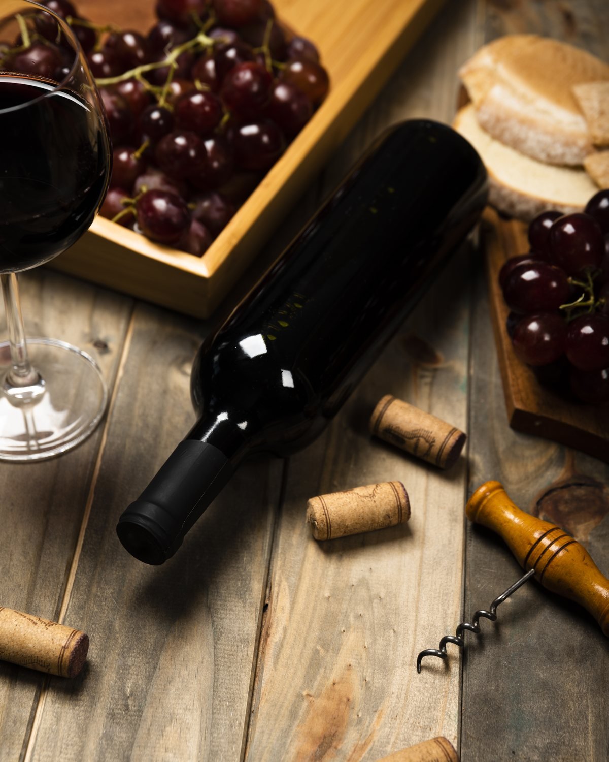 Botella de tinto tumbada sobre una mesa de madera, con una copa servida, racimos de uva y un sacacorchos